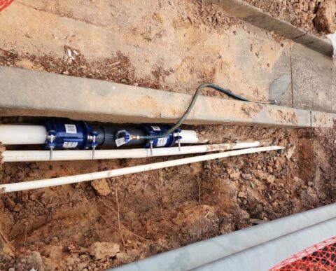commercial water supply line with 2 blue steel Hymax coupling and a new section of piping in the middle. There is a pressure measuring valve in the new piping and a hose for testing is attached. This, along with other pipes, is in a trench beside a highway guard rail.