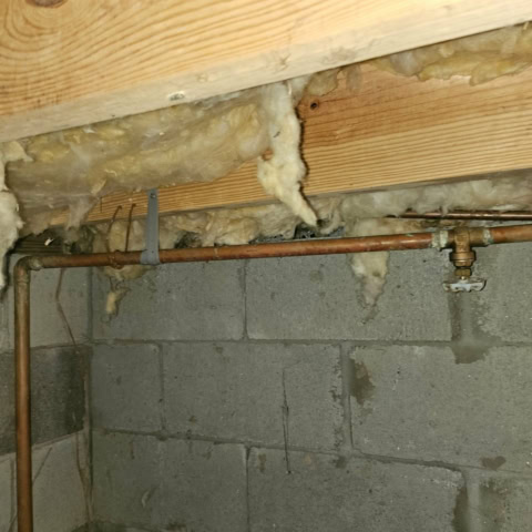 A water supply pipe in a ceiling with a valve with a red handle that can turn off the line. There are wooden beams and a cinderblock wall.