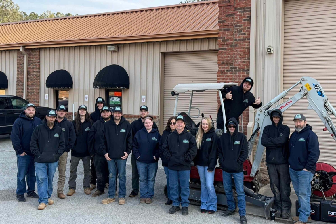 Reynalds Brothers Plumbing & Environmental Team Photo In Front of our Building Door, with Reynalds Brothers logo on the door, on all of the hats that cover everyone but 2 women, and on all of the sweatshirts that everyone is wearing. There is a biuding behind that is made of steel panel, has 1 door and 1 window and 2 garage doors, one in the forground and one closer of the right side. A mini excavator is also parked behind the team.