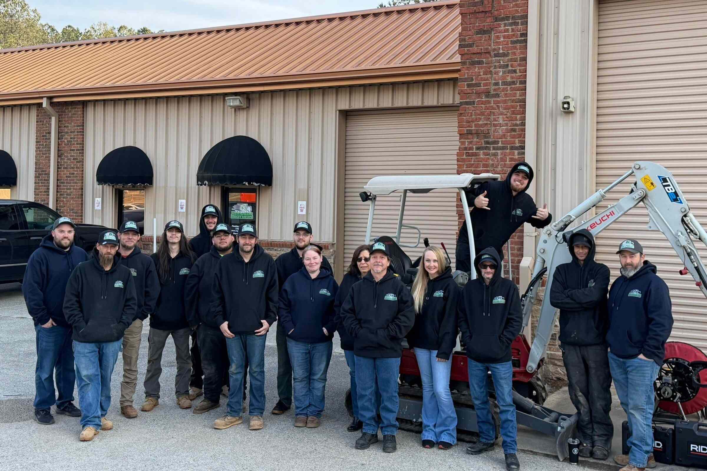 Reynalds Brothers Plumbing & Environmental Team Photo In Front of our Building Door, with Reynalds Brothers logo on the door, on all of the hats that cover everyone but 2 women, and on all of the sweatshirts that everyone is wearing. There is a biuding behind that is made of steel panel, has 1 door and 1 window and 2 garage doors, one in the forground and one closer of the right side. A mini excavator is also parked behind the team.