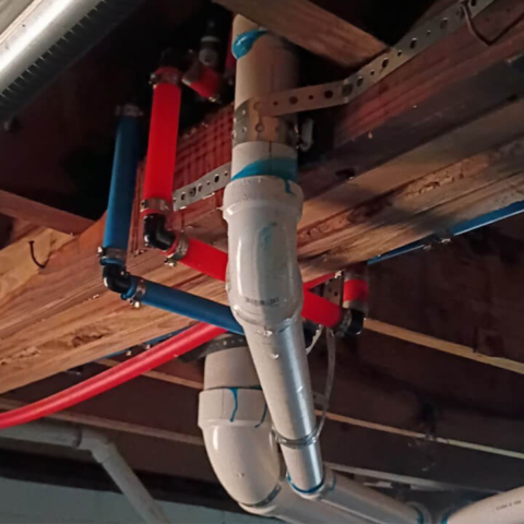 An upward view of exposed residential plumbing in an unfinished ceiling. Thick white PVC drain pipes and flexible red and blue PEX water supply lines are routed between wooden floor joists and secured with metal strapping.