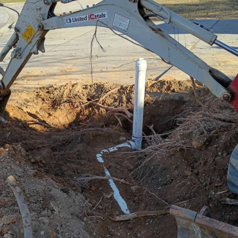 An excavated dirt hole showing a newly installed white PVC sewer line connecting to a tall vertical cleanout pipe. The arm of a construction excavator stretches over the hole, and exposed tree roots are visible in the dirt walls.