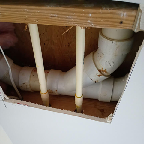 A square hole cut into white drywall revealing hidden plumbing. Large white PVC drain pipes and two thinner, yellowish water supply lines run through the wooden framing inside the wall or ceiling cavity.