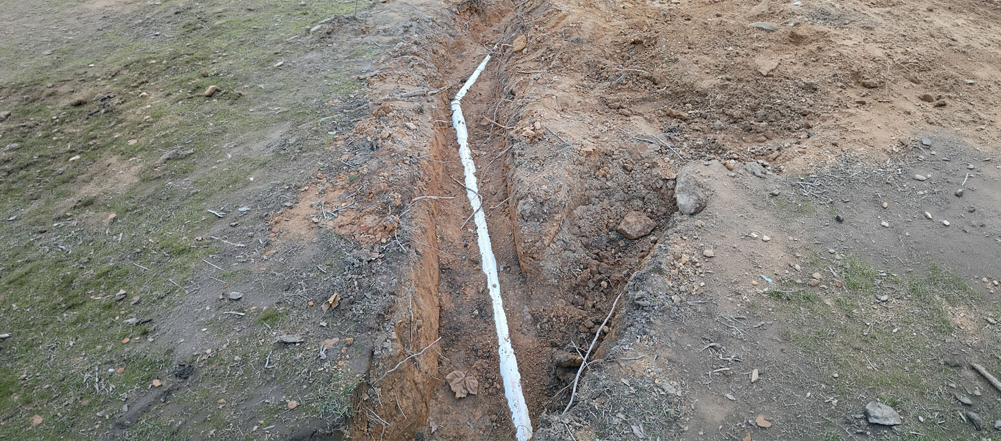 An exposed residential sewer line made of white PVC pipe laid in a freshly dug dirt trench.