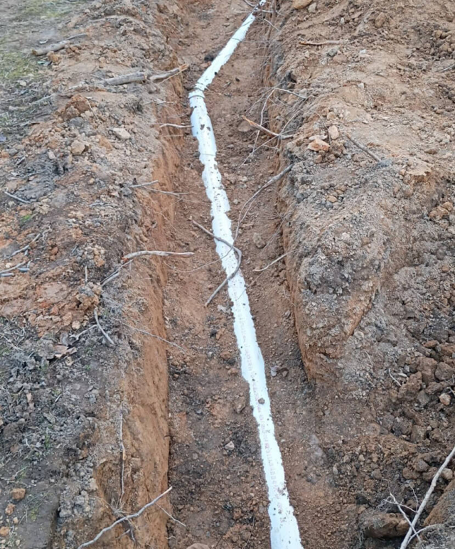 A long white PVC sewer pipe lying at the bottom of a freshly dug dirt trench.