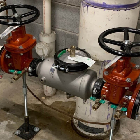 Picture shows a newly installed back-flows prevention assembly on a large industrial water main inside a facility. There is a grey cinder-block wall behind, concrete floor beneath, and a fenced off area on the right side of the phone. The piping descends from the ceiling, turns 90 degrees to run parallel to the ground for 4 feet, around 2 feet high, and then ascends to the ceiling again, and a new back-flow prevention assembly has been installed in that parallel part of the piping.