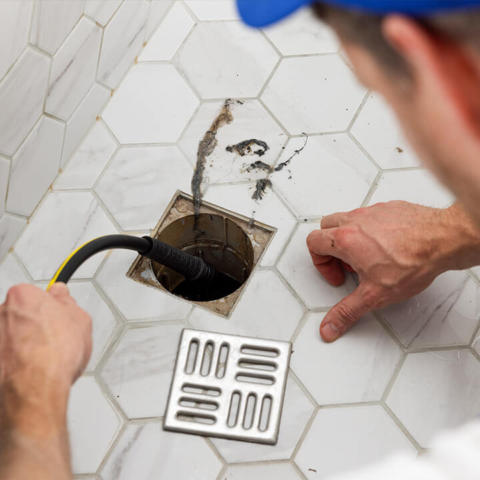 A professional plumber uses a cable auger to clear a hair blockage from a tiled shower drain, showcasing effective residential drain cleaning services.