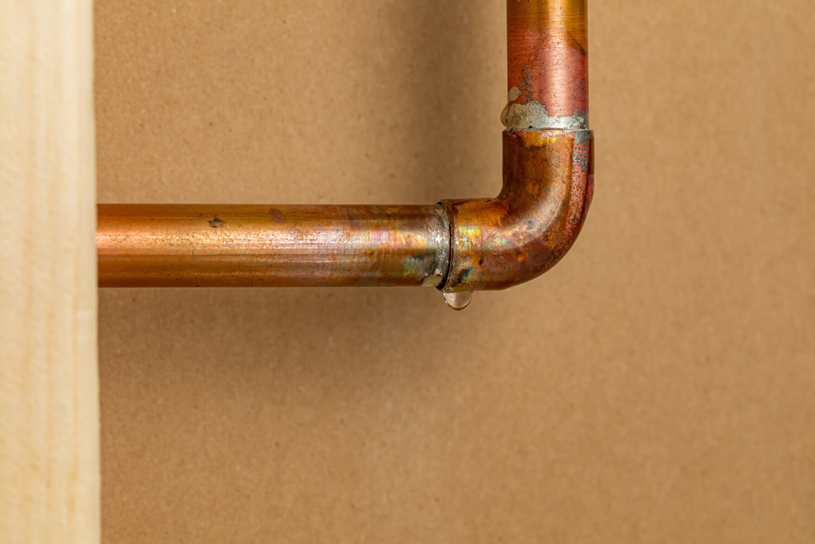 A close-up of a copper pipe elbow with a visible water droplet leaking from a joint, highlighting the need for professional pipe repair and leak detection services.