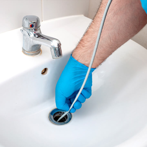 A plumber wearing protective gloves uses a professional drain snake to clear a stubborn clog from a bathroom sink, demonstrating effective residential drain cleaning services.