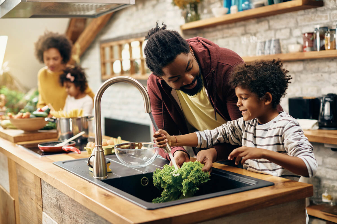 A father and son use a modern pull-down kitchen faucet to wash vegetables, illustrating the importance of reliable residential plumbing for everyday family activities and home health.