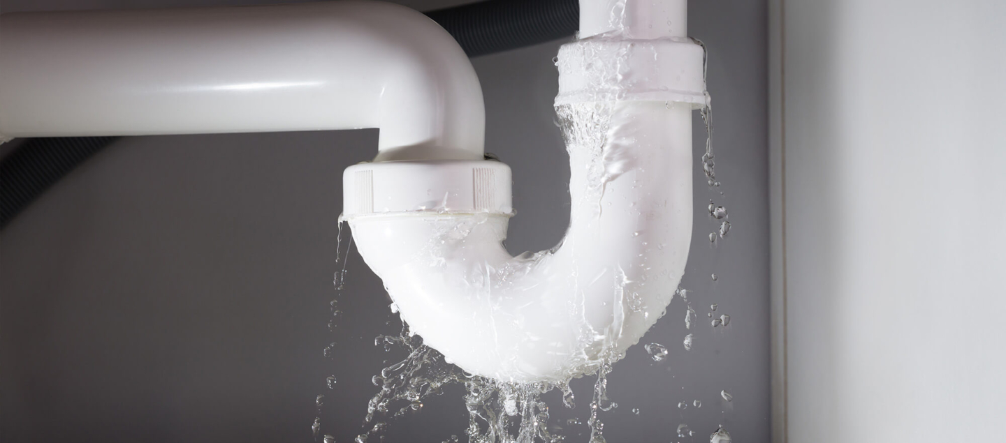 A white PVC P-trap under a sink with significant water spraying from the joints, emphasizing the urgency of professional emergency plumbing repair services.